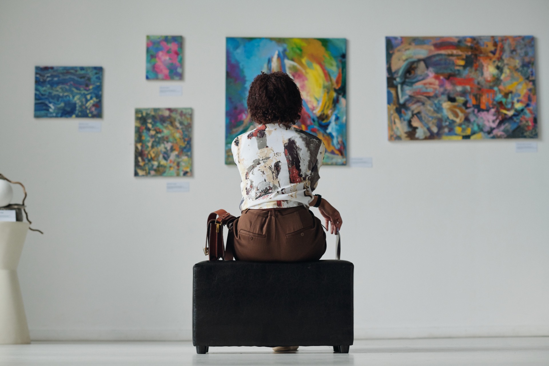 A woman sitting on a bench in an art gallery, looking at several colorful abstract paintings displayed on the wall.